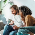 A couple sit on a sofa and smile at a laptop they look at together. 